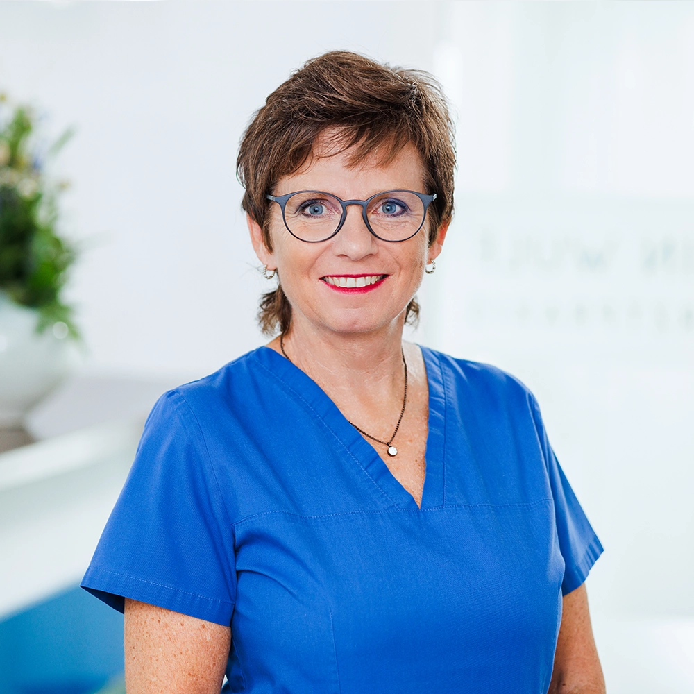 Portrait Praxisteam Gisela Mitarbeiterin Gisela mit Kurzhaarfrisur und Brille lächelt in blauer Praxiskleidung